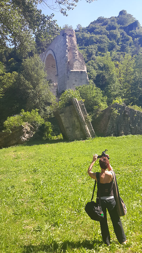 Woman standing in grassy field filming an ancient arch in the distance