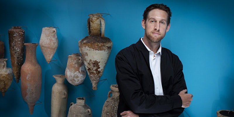 A photograph of Emlyn Dodd, a white man in a black jacket standing in front of Roman pottery 