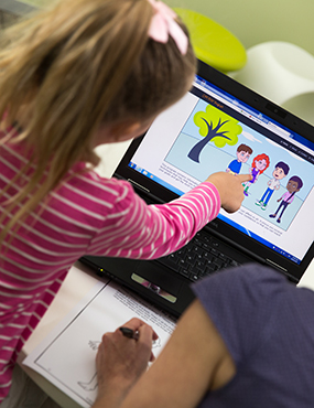 A child and an adult are accessing the Cool Kids program on a laptop. The child is pointing at the screen, which shows several cartoon people.
