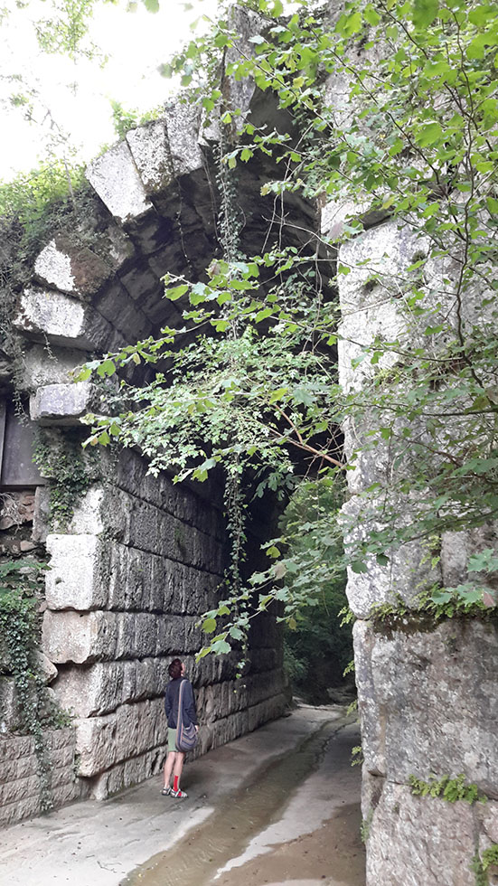 Woman standing underneath massive ancient stone arch overgrown with plants