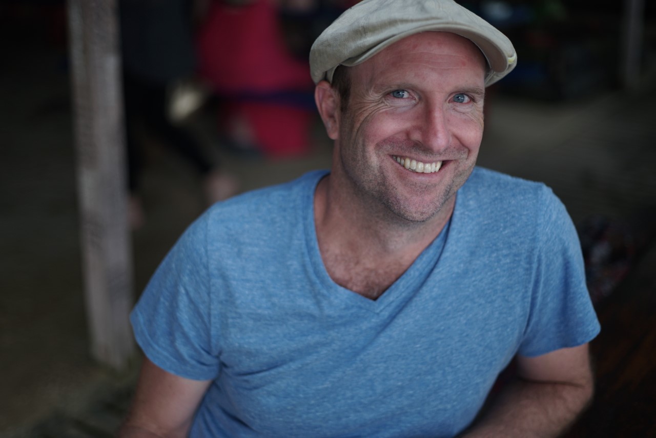 A photograph of Tom smiling at the camera. The background is blurred