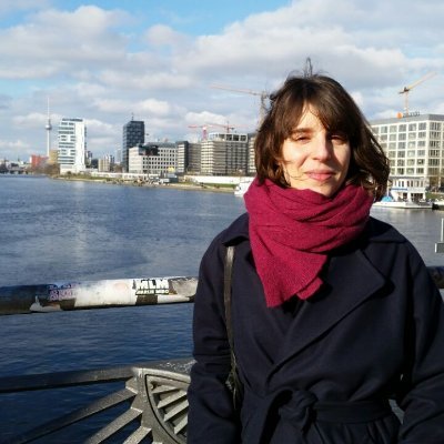 An Image of Dr Camilla Di Biase-Dyson: a woman with shoulder length brown hair, wearing a red scarf and a black jacket. She stands to the right of the image, and in the background we see a blue waterway.