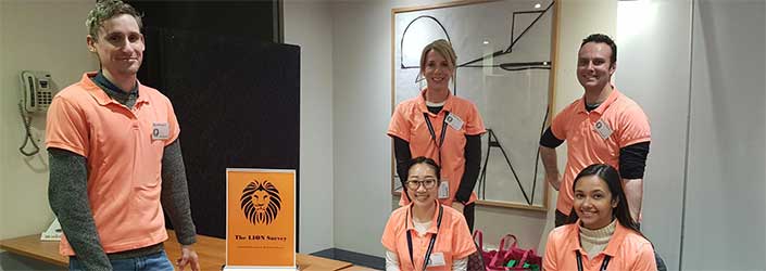 Five people in orange polo shirts around a table with materials encouraging participation in the Lion survey for the Ethos program.