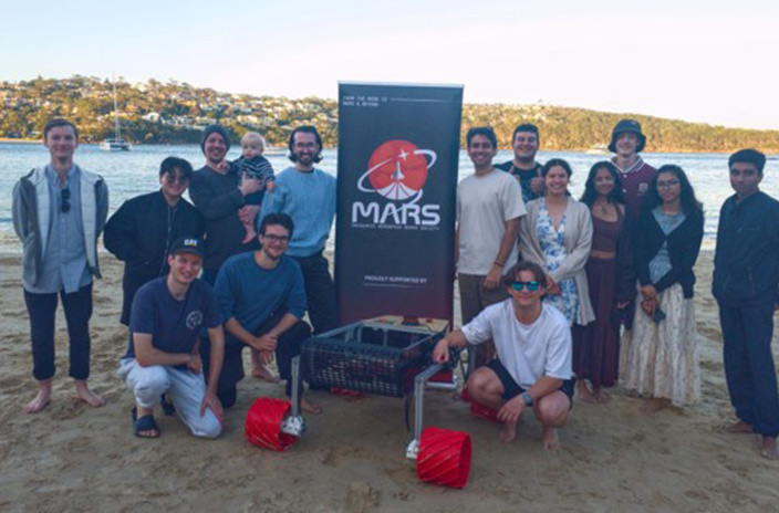 The Macquarie Aerospace Rover Society (MARS) team standing on a beach with Wally the rover.