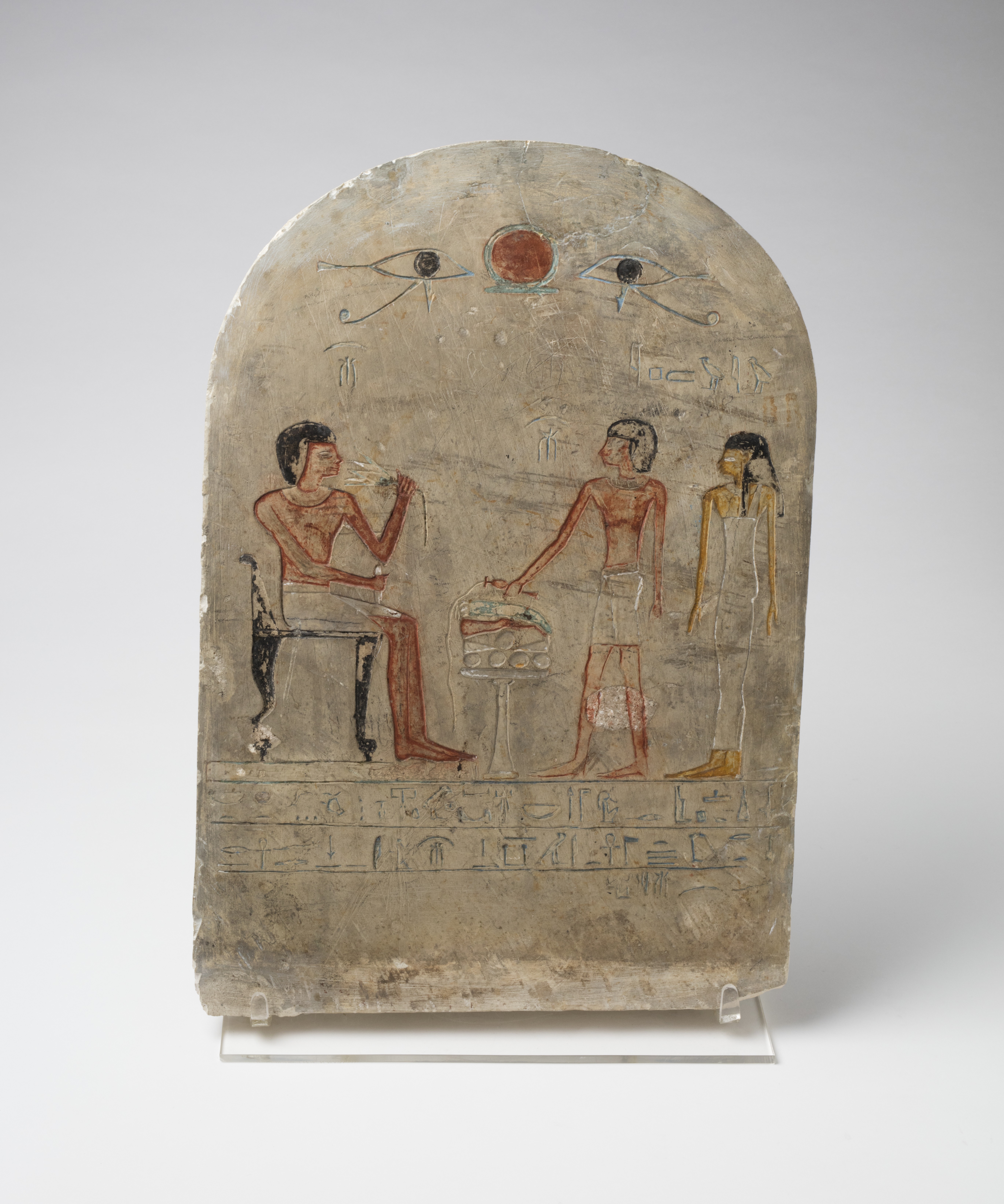 An ancient Egyptian painted stone stelae. A man is seated at a table of food offerings. Two people, a man and a women are presenting him gifts. there is hieroglyphic text below this painting.