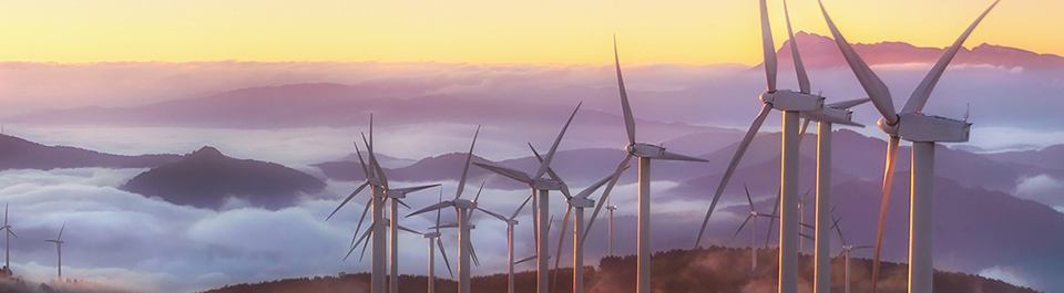 A cluster of wind turbines on a hilltop at dawn. Several hills break through the sea of mist like islands. The rising sun colours the sky pale yellow and the mist pale purple.