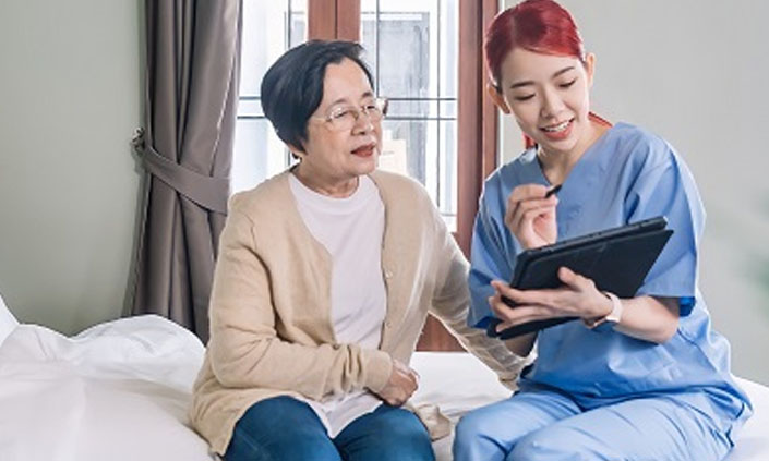 An elderly patient and a nurse sitting on a bed looking at an iPad.