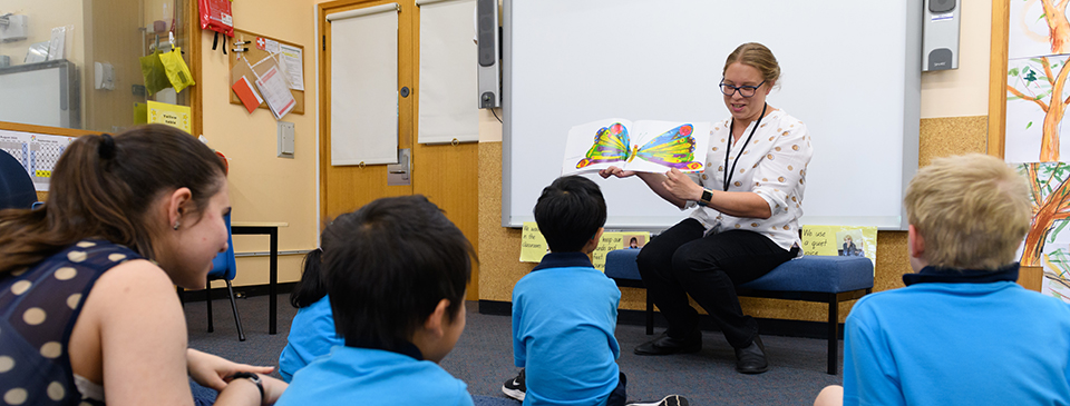 A teacher sits in front of a SMART board, holding a copy of The Very Hungry Caterpillar open near the end. Another teacher and four children in blue shirts sit cross-legged on the floor in front of her.