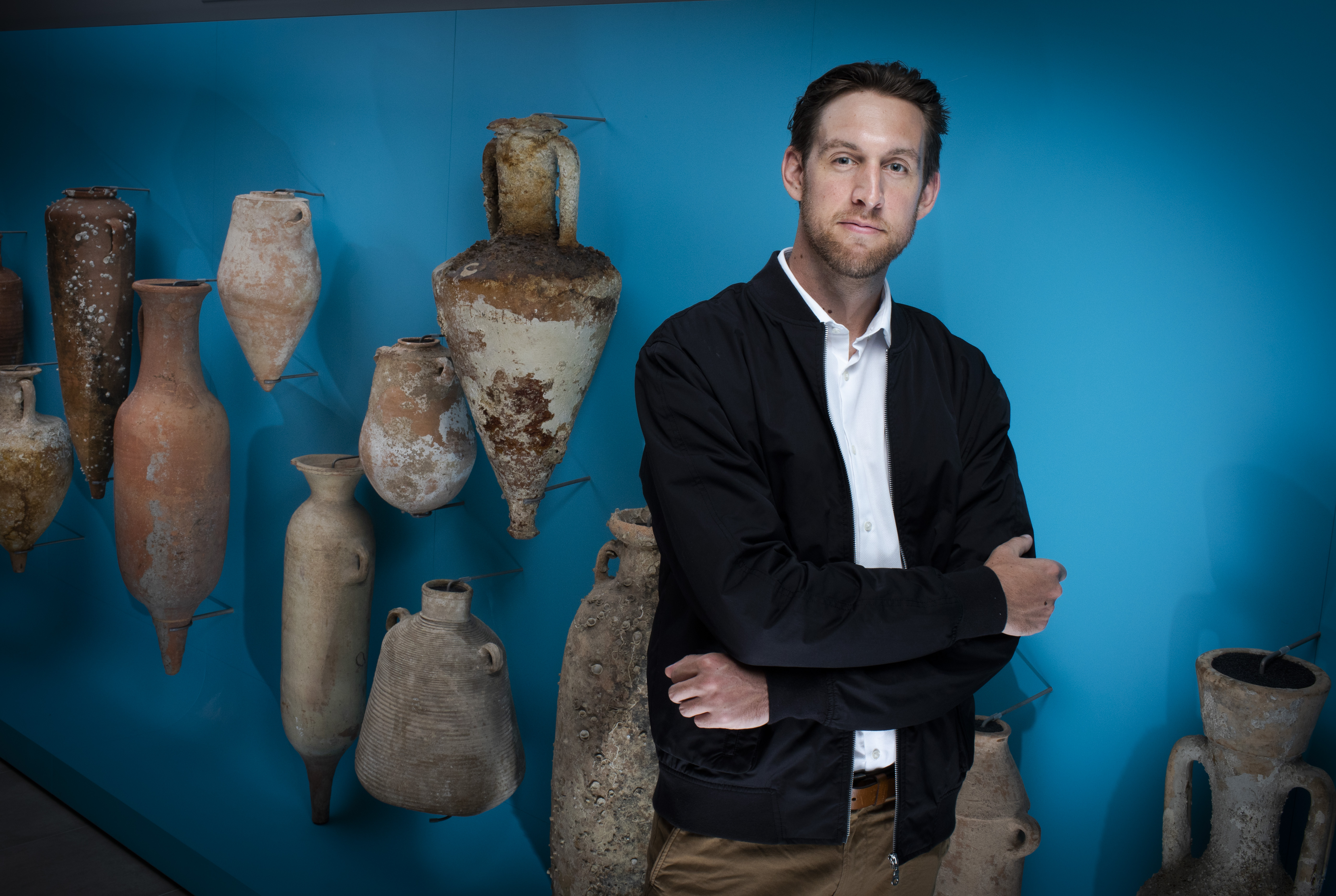 Photo of Emlyn Dodd standing in front of blue wall displaying ancient jars