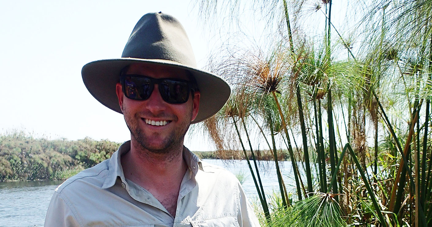 A photograph of a white man in front of a river with a large brim hat and sunglasses on