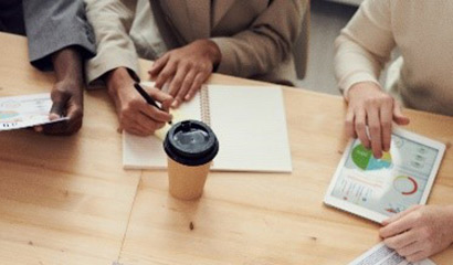Three people sitting at a table. One holds a piece of paper, another has a coffee cup and an open notebook in front of them, and the last is using a tablet to look at graphs and charts.