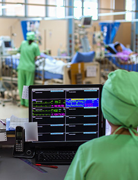 An intensive care unit with several beds, read-outs on a screen, and two medical professionals in full PPE.