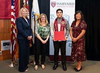 Professor Westbrook with a graduate and two other people in front of the Harvard Medical School banner
