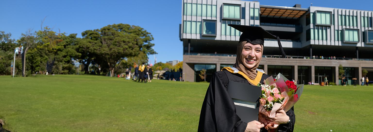 Image of a girl in her graduation gown holding flowers and standing on the green lawn on campus