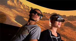 Two people with VR goggles in front of projected image of desert