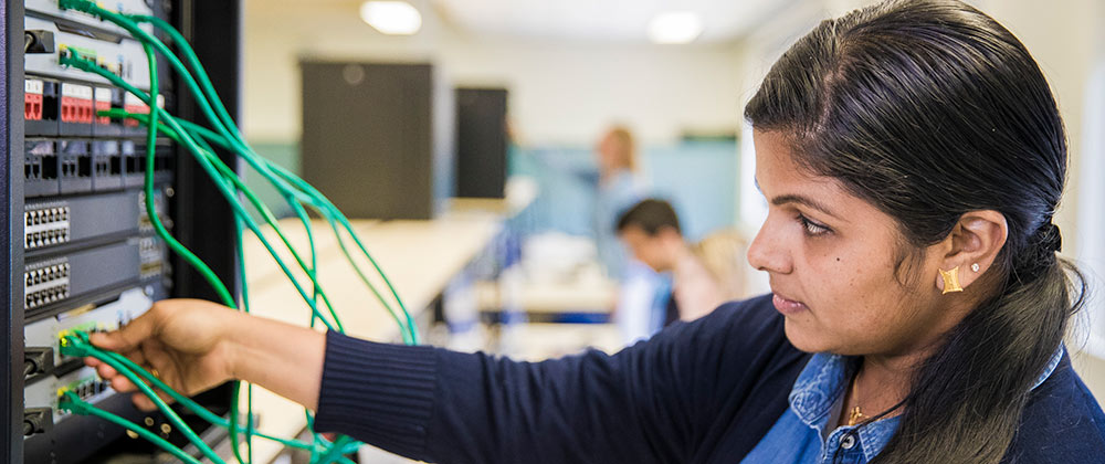 A student in a computer lab, connecting green cables to the back of a server.