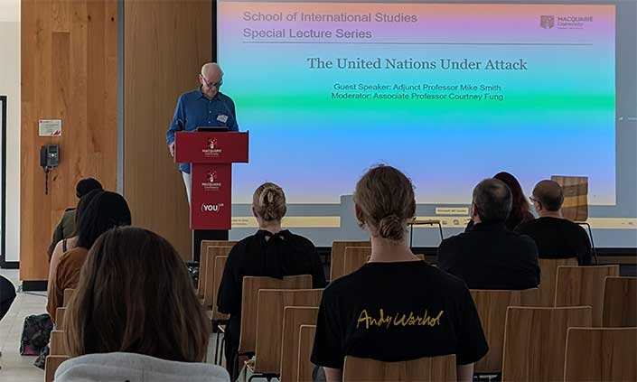 Adjunct Professor Mike Smith talking to a room of people with a screen behind him that says 'The United Nations Under Attack'