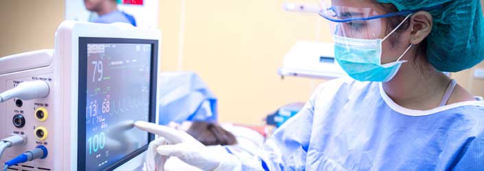 A person wearing scrubs, a hair net and a face mask pointing at a hospital screen with a patient laying on a table in the background.