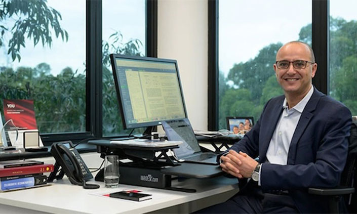 Professor Dali Kaafar smiling and sitting at desk in suit