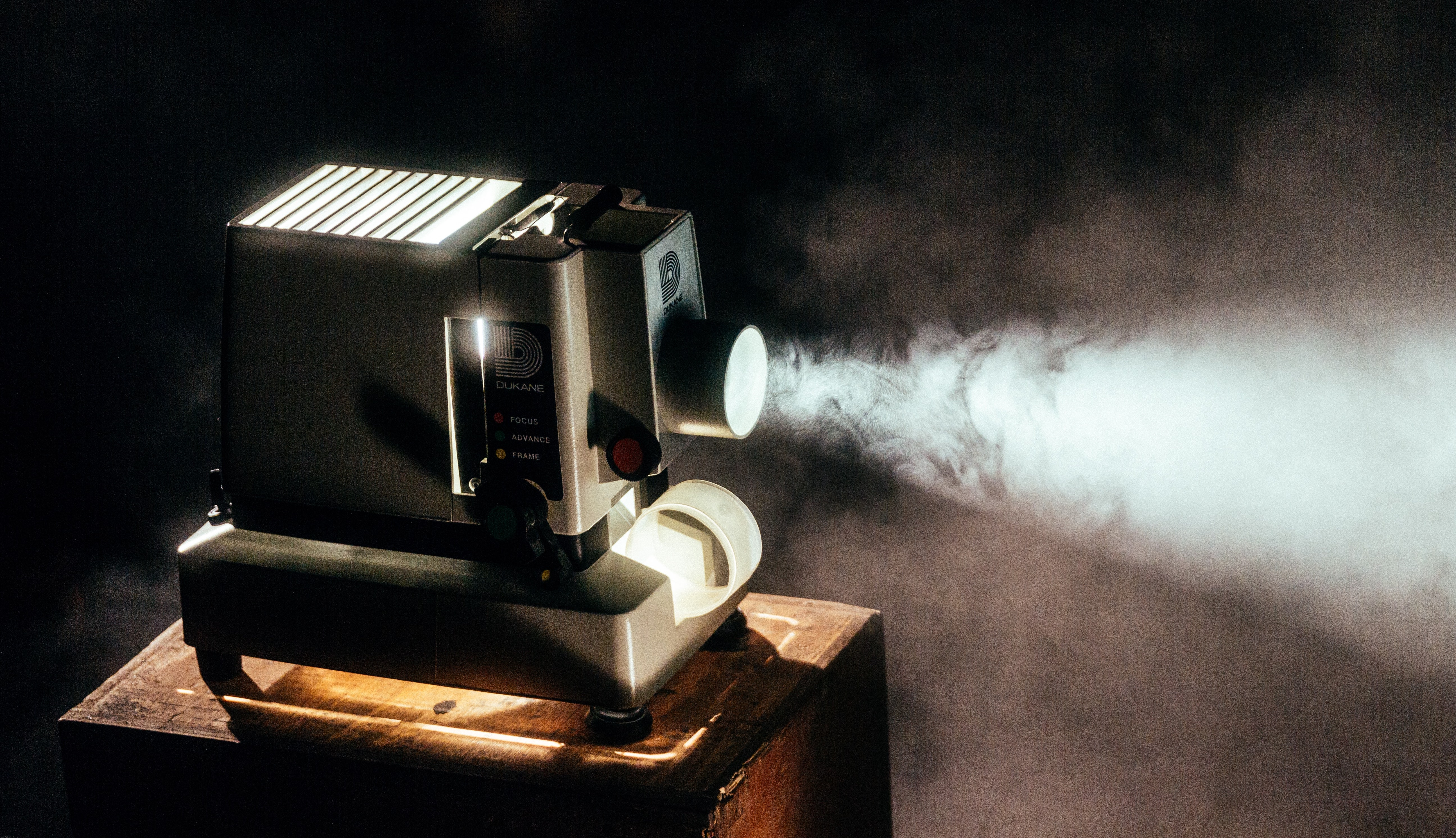 An old projector sits on a wooden table and shines through a dark room. The image that is being projected is not shown.