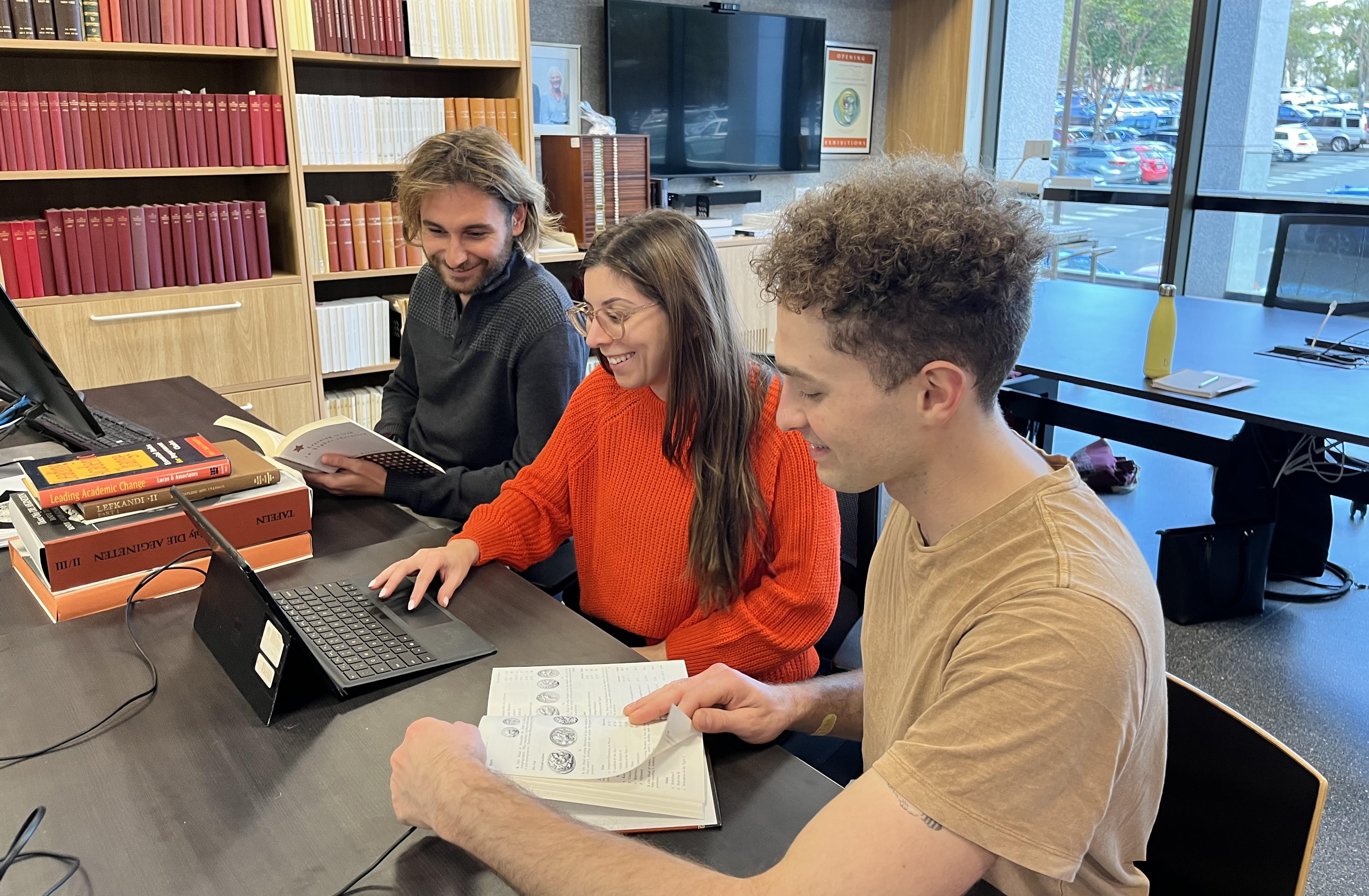 Three people sitting at ACANS workstation looking at laptop and book