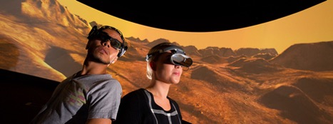 Two people wearing virtual reality goggles in front of projection of desert landscape