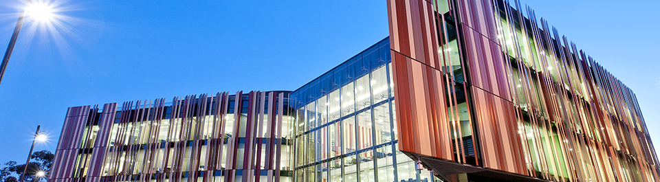 The front of the Library building at dusk with brightly lit windows and the iconic rust and sand coloured panels contrasting against the deep blue sky.