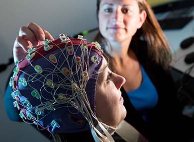 A person wearing a brain examination cap with a woman adjusting one of the nodes.