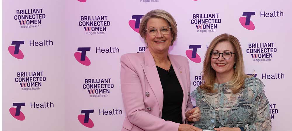 Professor Johanna Westbrook receiving an award from a woman. They are standing in front of a backdrop of logos for Brilliant Connected Women and Telstra Health
