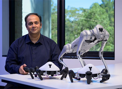 Dr Adnan Mahmood sitting and desk holding controller with three robots on a white desk