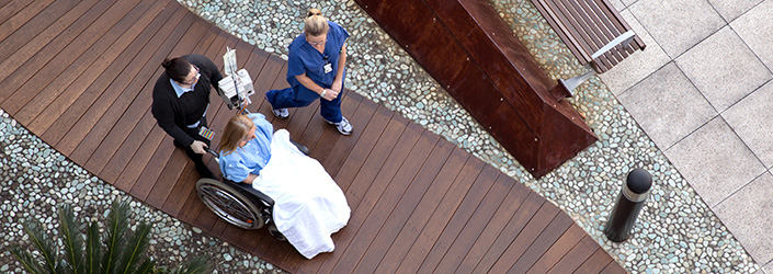 Nurses walk patient in wheelchair along wood-planked courtyard.