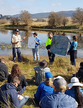 The River Styles short course in Mulwaree. Instructors stand in front of a river, holding a whiteboard and a map up for the students seated nearby to see.