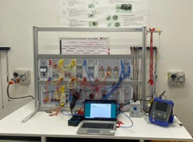 An electric vehicle charging station in construction on top of a workbench. There are multicoloured wires running across it, connecting the various switches and circuits, and various monitoring devices connected to it, including a laptop.