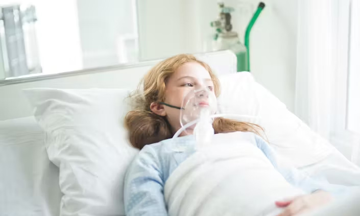 A young person in a hospital bed wearing an oxygen mask.