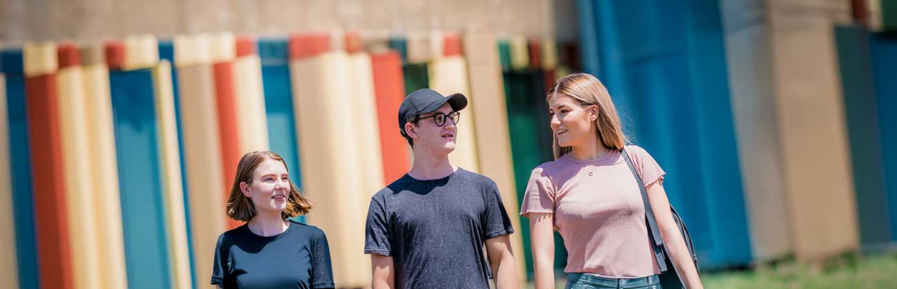 Three higher degree research students walk together in front of the Library building's iconic rust and sand panels.