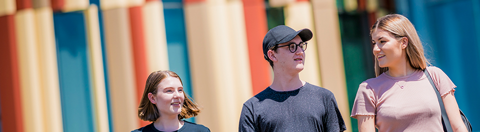 Three higher degree research students walk together in front of the Library building's iconic rust and sand panels.