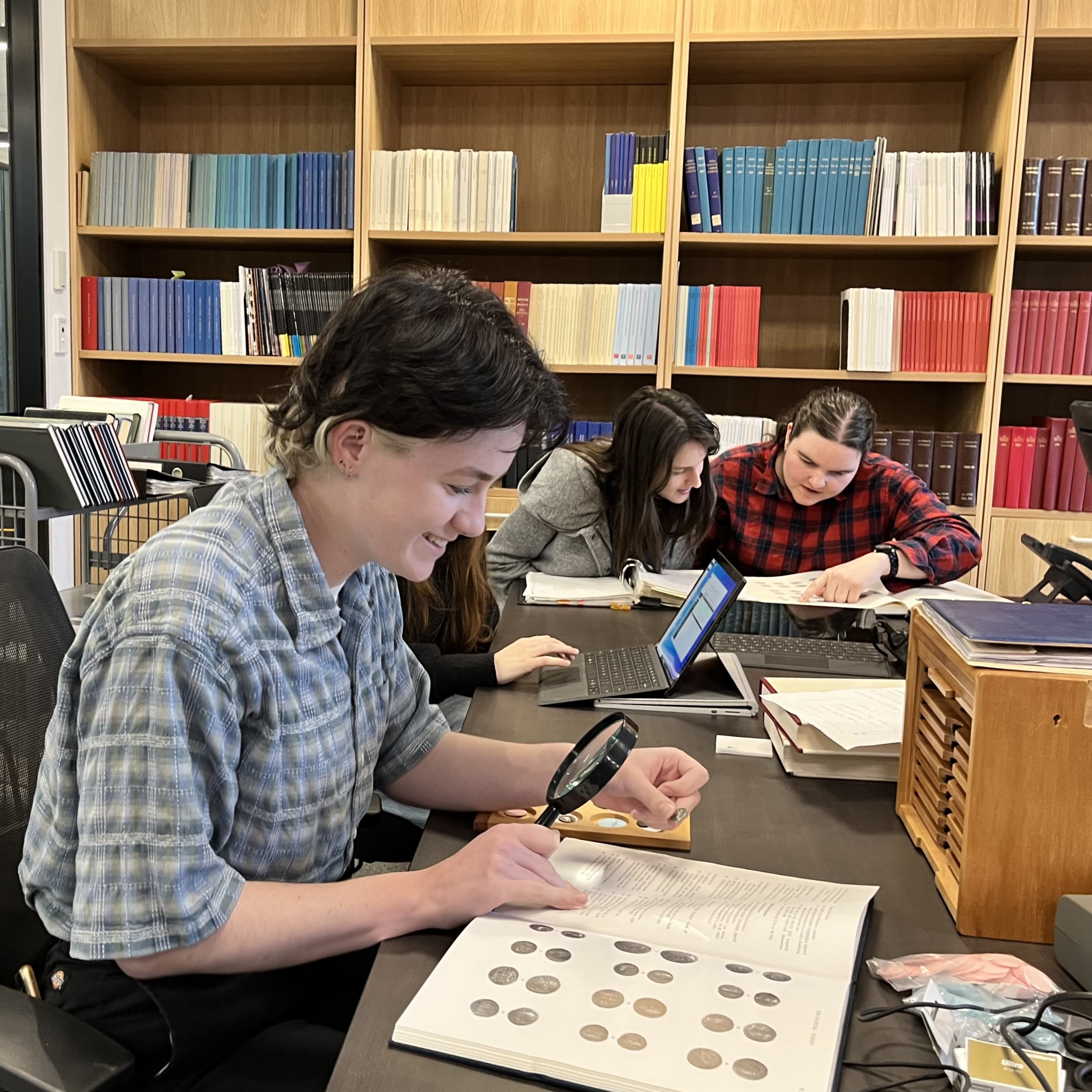 Image of a smiling person looking thrugh a magnifying glass at some coins, two other people in background