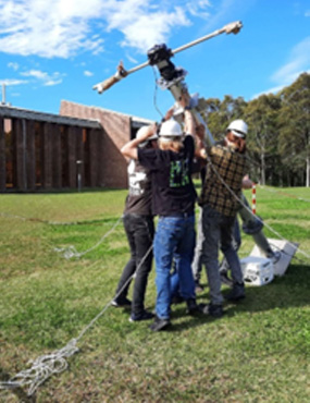 Three people in hardhats outside on the lawn lifting a tall metal object.