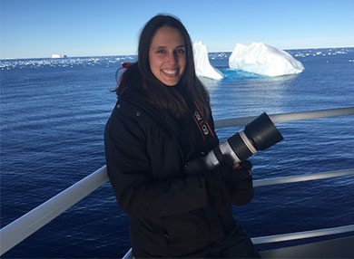 Dr Vanessa Pirotta posing holding a camera on a boat in front of icebergs