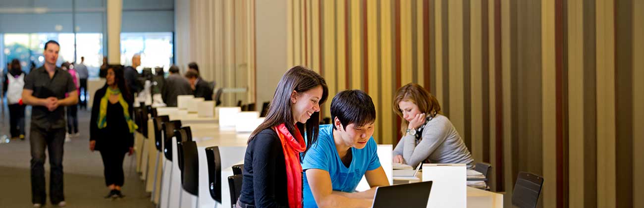 Three students sit on high stools at a long bench near the Library entrance, looking at books and laptop screens.