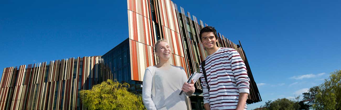 Two students holding bags and books stand in front of the Library building's iconic rust and sand panels.
