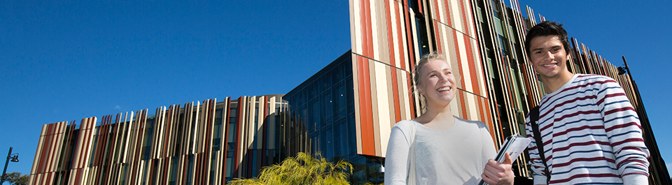 Two students holding bags and books stand in front of the Library building's iconic rust and sand panels.