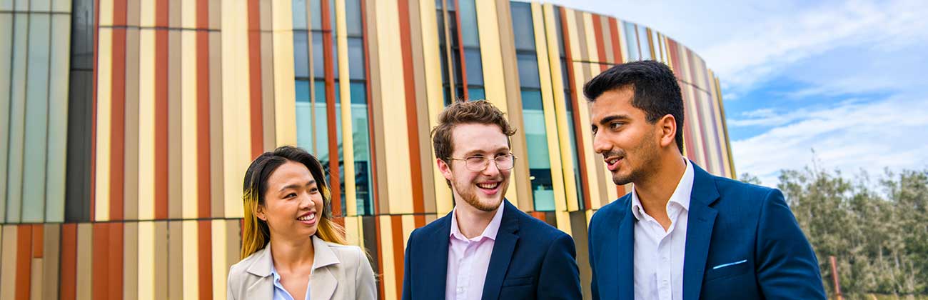 Three young professionals in smart casual attire sit talking in front of the Library building's iconic rust and sand panels.
