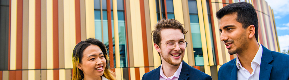 Three young professionals in smart casual attire sit talking in front of the Library building's iconic rust and sand panels.