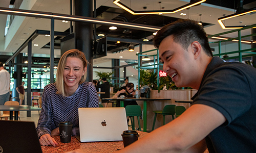 Two students sitting at a table in the Central Courtyard Hub, smiling and looking at a laptop screen.
