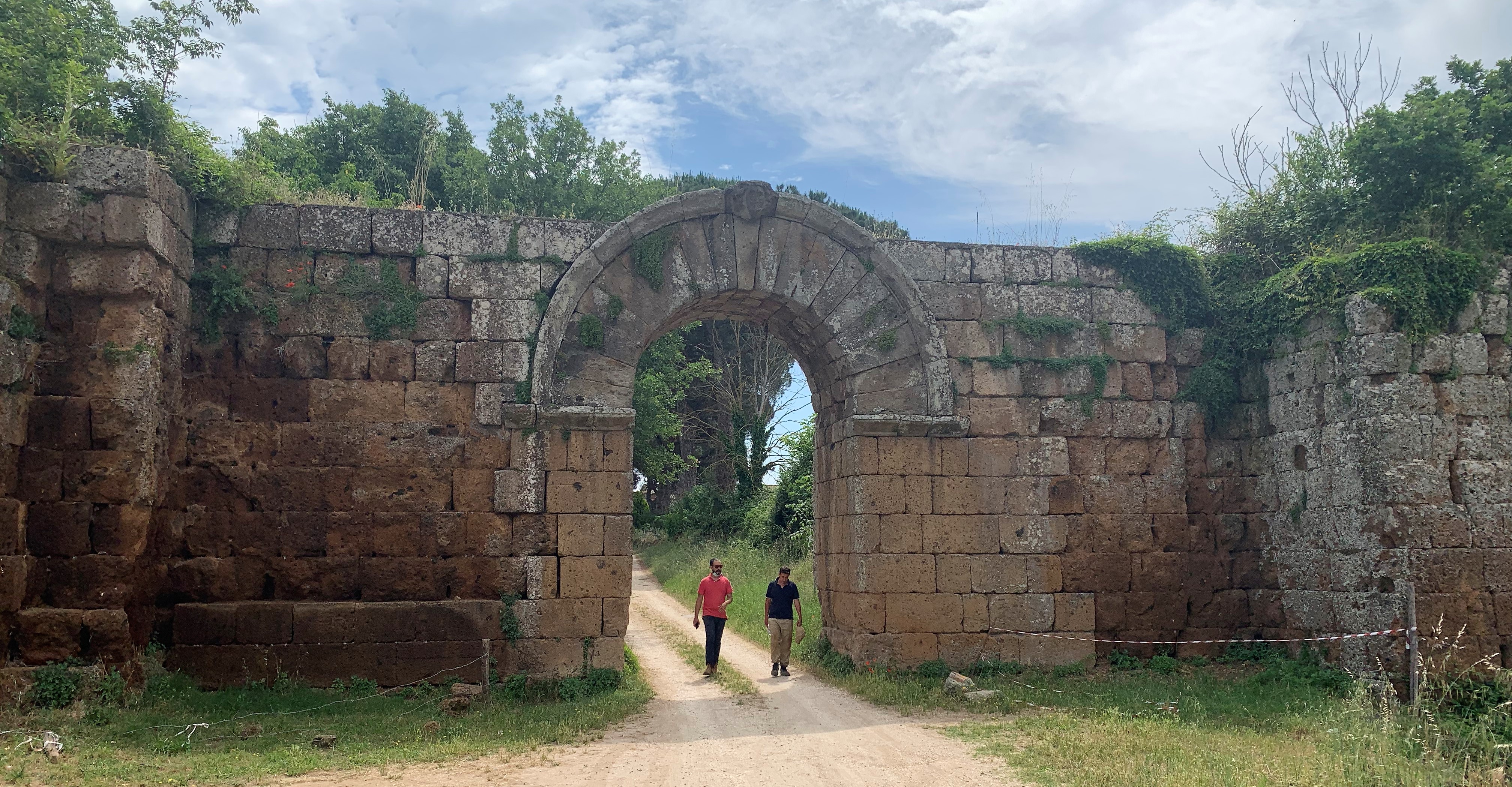 A photograph of a large roman gate with two people walking through