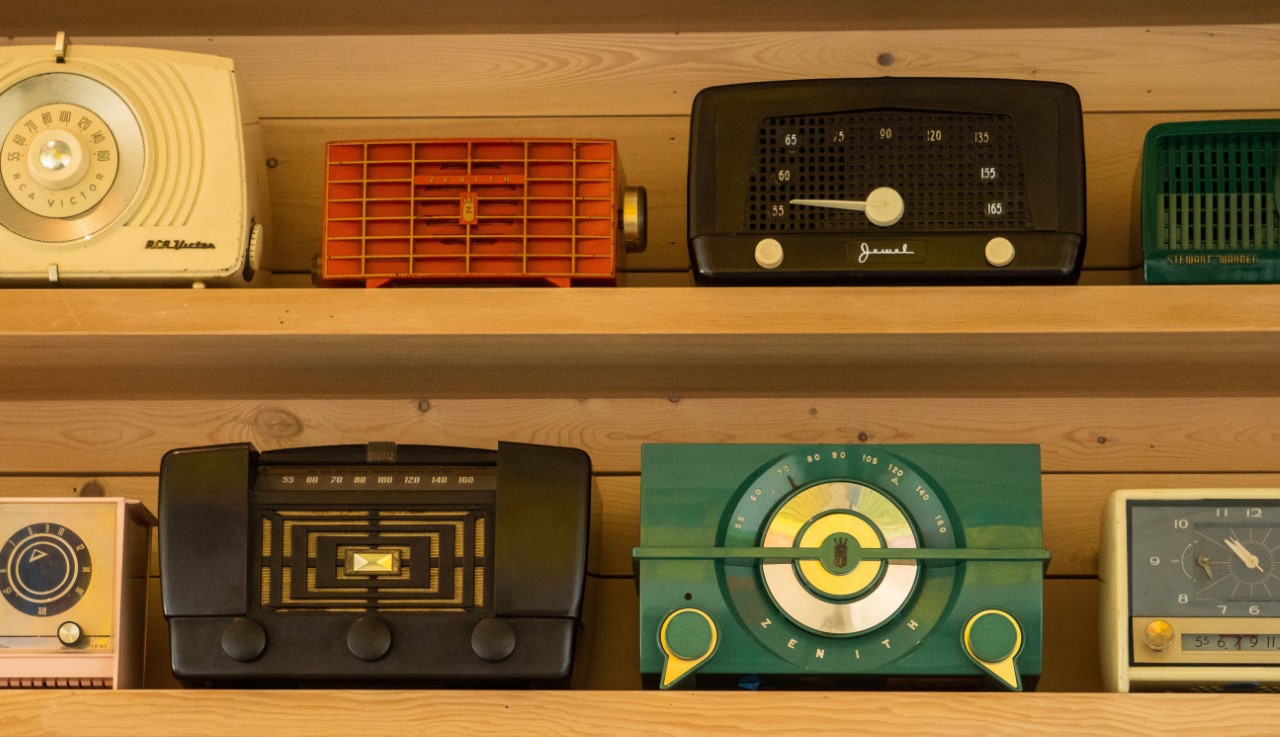 Two wooden shelves, each holding four older radios. These radios are cream, orange, black and teal.