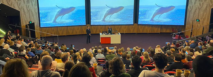 Photo from back of full lecture room, lecturer at lectern presenting with humpback whales on the screens behind 