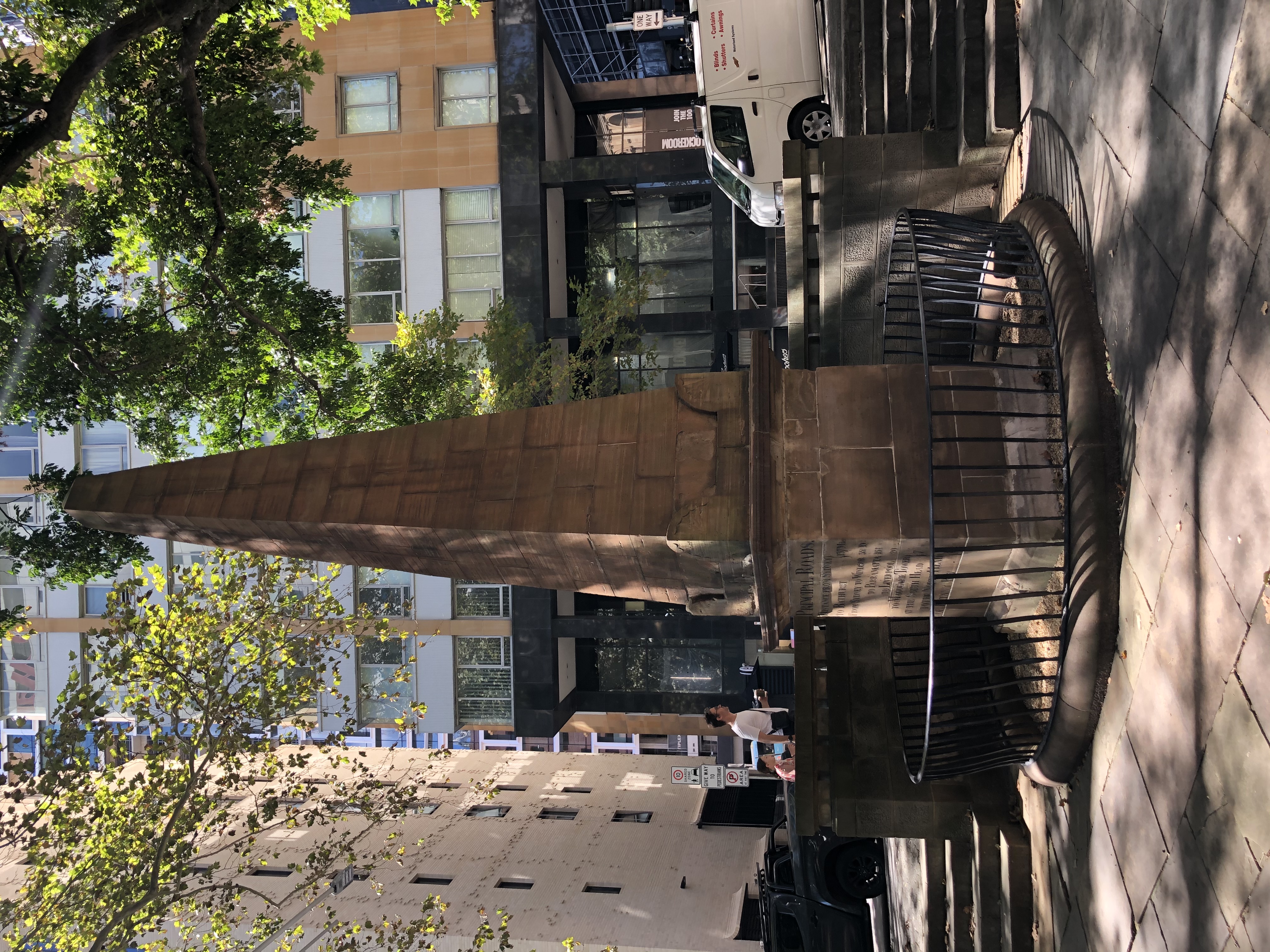 A photograph of an obelisk in the streets of Sydney, made of stone with writing carved.  