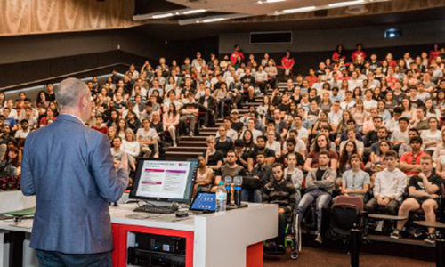 A lecturer speaks to a large lecture theatre full of students.